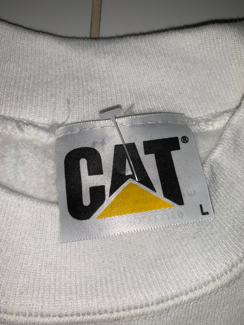 Pu&ograve; includere: Felpa bianca con un'etichetta logo CAT nera e gialla. L'etichetta ha la parola "CAT" in lettere nere con un triangolo giallo sotto. L'etichetta ha anche un'etichetta di taglia che dice "L".
