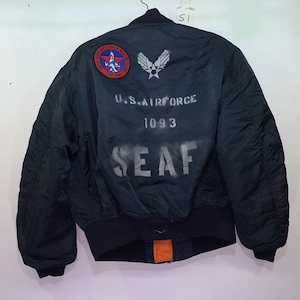 Könnte beinhalten: Marineblaue Bomberjacke mit schwarzem geripptem Kragen und Bündchen. Auf der Rückseite der Jacke befindet sich ein weißer Schablonentext mit der Aufschrift "U.S. AIR FORCE 1093 SEAF". Auf dem oberen Rücken befinden sich Aufnäher mit Text und Emblemen.