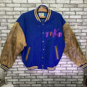 May include: A blue and brown varsity jacket with leather sleeves. The jacket has a gold and white striped collar and cuffs. The word "TOKO" is embroidered on the front in purple.