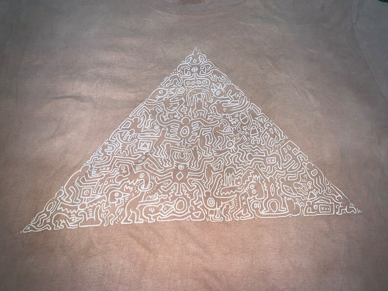 May include: A beige t-shirt with a white outline of a pyramid filled with abstract figures and symbols.