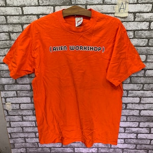 May include: Orange t-shirt with the text "(ALIEN WORKSHOP)" printed in black.