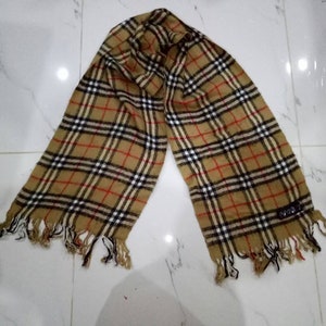 May include: A brown, black, and red plaid scarf with fringe on the ends. The scarf has a small black tag with the word "SPIGA" on it.