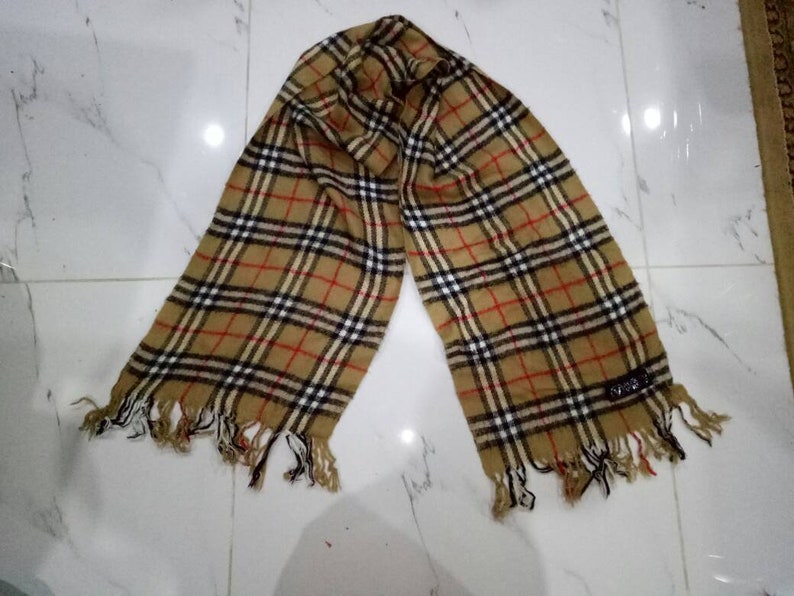 May include: A brown, black, and red plaid scarf with fringe on the ends. The scarf has a small black tag with the word "SPIGA" on it.