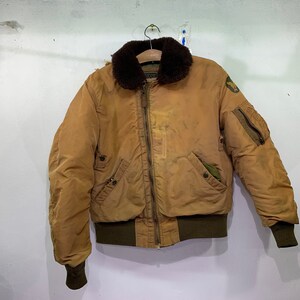 Puede incluir: Una chaqueta bomber marrón con cuello de piel sintética y cierre de cremallera. La chaqueta tiene dos bolsillos en la parte delantera y un parche en la manga izquierda.