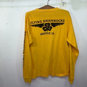 May include: Yellow long-sleeve shirt with black text and a graphic of a winged shamrock. The text reads "Flying Shamrocks MC Norfolk, VA."