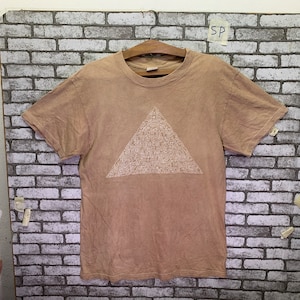 May include: A light brown t-shirt with a white triangle design. The triangle is filled with a pattern of small white shapes.