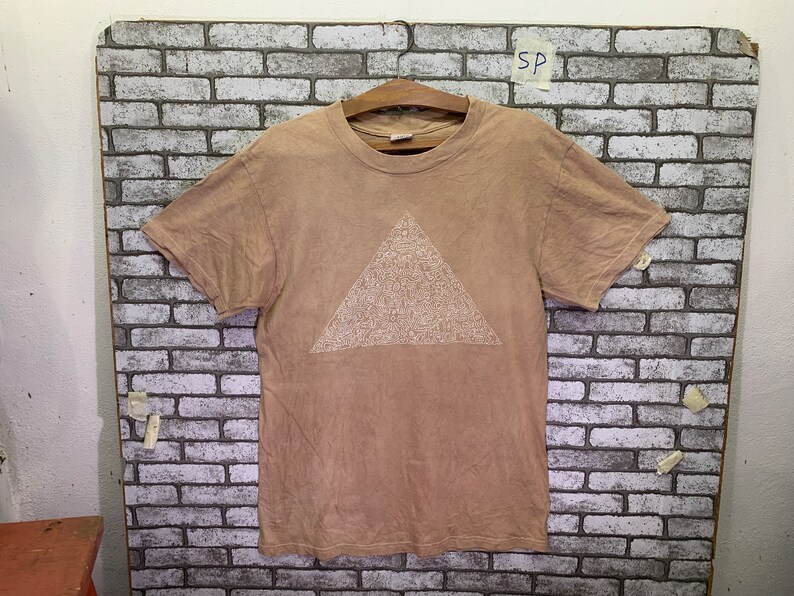 May include: A light brown t-shirt with a white triangle design. The triangle is filled with a pattern of small white shapes.
