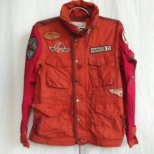 Könnte beinhalten: Eine rote und burgunderfarbene Jacke mit mehreren Patches, darunter "HANGER 75" auf der Vorderseite. Die Jacke hat einen Knopfverschluss und zwei große Taschen auf der Vorderseite.