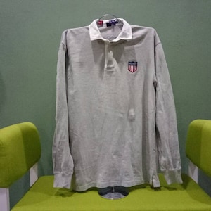 Puede incluir: Una camisa polo de manga larga de color gris claro con un pequeño logotipo de escudo rojo, blanco y azul en el pecho izquierdo. La camisa tiene un cuello blanco y una tapeta de botones.