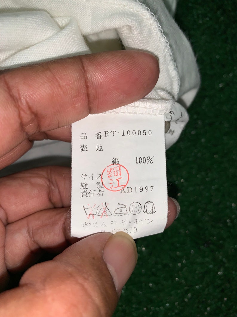 Pu&ograve; includere: Un'etichetta bianca con testo nero e un timbro rosso. L'etichetta dice "品番RT-100050 表地 鍋 100% サイズ細 縫製江 責任者 AD1997".