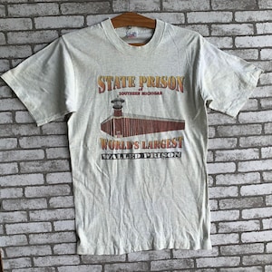 May include: A light gray t-shirt with a graphic print of a prison building. The text on the shirt reads "State Prison Southern Michigan World's Largest Walled Prison".