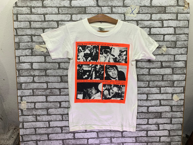 May include: White t-shirt with a black and white graphic print of a band. The graphic print has the text "Being the adventures of a young man... who loved a bit of the old ultra violence went to jail, was brainwashed... and came out cured...or was he?"