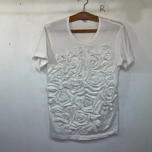 Pu&ograve; includere: Maglietta bianca a maniche corte con un design floreale 3D sul davanti.