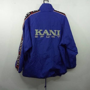 May include: A blue windbreaker jacket with the word "KANI SPORT" embroidered on the back. The jacket has black and red stripes down the sleeves with the word "KANI" repeated on the stripes.