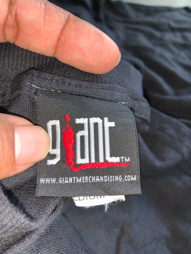 May include: Black tag with white text that reads "giant" with a red stick figure logo. The text "www.giantmerchandising.com" is below the logo. The tag has a white label with the word "medium" on it.