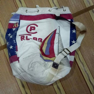 May include: A white canvas drawstring bag with a red, white, and blue American flag design. The bag features a sailboat graphic and the text "RL-93" and a circled "P".