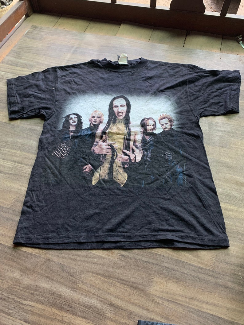 May include: Black t-shirt with a graphic print of a band. The band members are wearing black clothing and have long hair. The band's name is "Marilyn Manson".