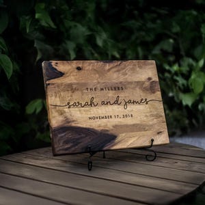 May include: A rectangular wooden cutting board with natural dark and light wood grain patterns. The board is engraved with the text THE MILLERS, sarah and james, and NOVEMBER 17, 2018. It is displayed on a black metal stand for upright viewing.