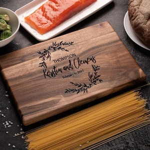 May include: A rectangular dark brown wooden cutting board featuring a laser-engraved design of a leafy wreath surrounding the text "THOMPSON", "Kerstin and Clemens", and "February 6, 2017" centered on the smooth, grained surface of the wood.