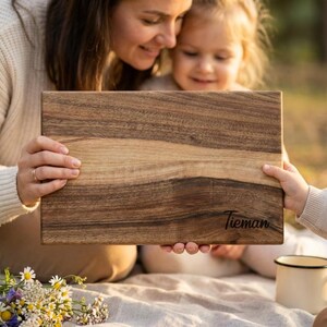 A woman and a young girl are sitting on a blanket in a park, with the woman holding a wooden cutting board with the name "Tieman" written on it.