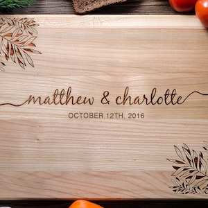 May include: A rectangular light-colored wooden cutting board featuring laser-engraved botanical leaf designs in the top-left and bottom-right corners. The center displays the text "matthew & charlotte" in script and "OCTOBER 12TH, 2016" below.