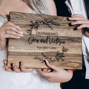 May include: A rectangular wooden cutting board featuring a dark wood grain pattern. The board is engraved with a floral wreath design, the name OKHRIMENKO, the names Den and Victoria in script, and the date September 7, 2018, centered on the front.