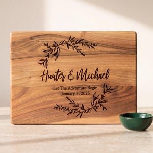 A wooden cutting board with a decorative wreath and the names Hunter & Michael printed on it, along with a small green bowl.