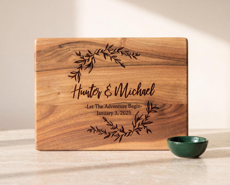 A wooden cutting board with a decorative wreath and the names Hunter & Michael printed on it, along with a small green bowl.