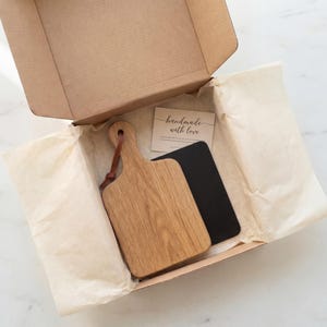 An open cardboard box containing a wooden cutting board and a black notebook, with a handwritten note inside.