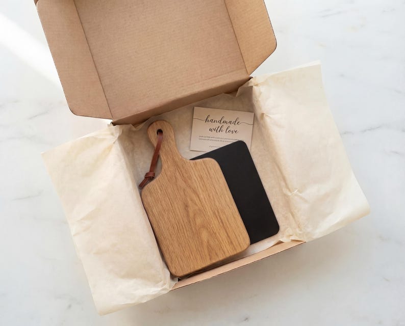 An open cardboard box containing a wooden cutting board and a black notebook, with a handwritten note inside.