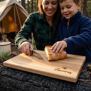 A woman and a young boy are preparing food together, with the woman cutting bread on a wooden cutting board. The cutting board has the name "The Luciers" engraved on it.