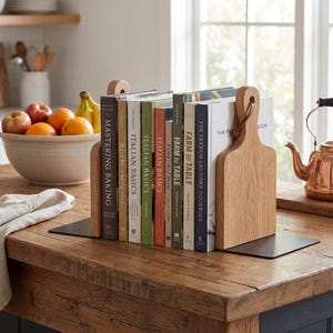 A collection of cookbooks is neatly arranged on a wooden table, with a bowl of fruit and a copper kettle in the background.