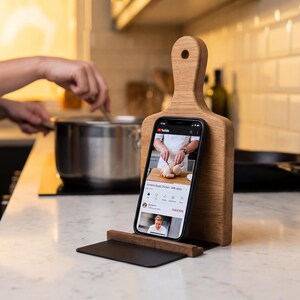 A person is cooking in a kitchen, with a smartphone displaying a cooking video on a wooden stand.