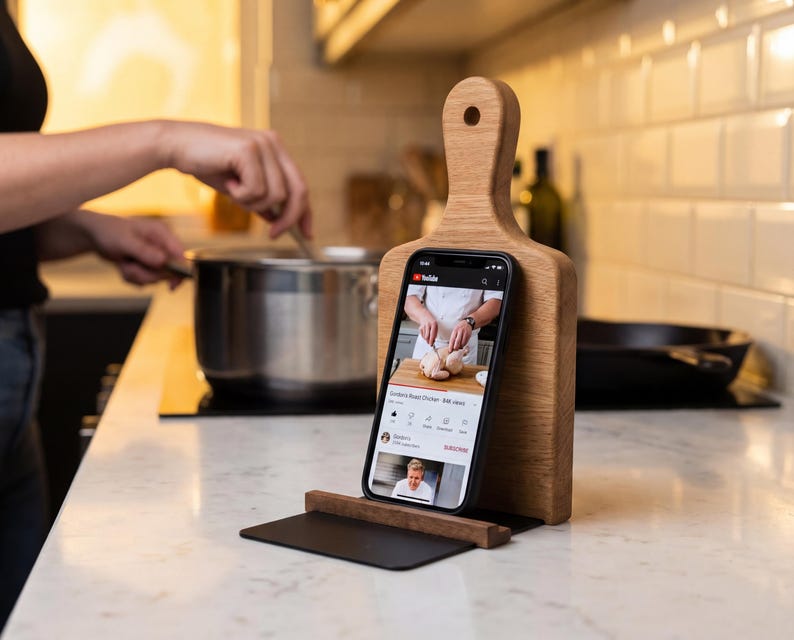 A person is cooking in a kitchen, with a smartphone displaying a cooking video on a wooden stand.