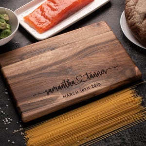 May include: A rectangular dark brown wooden cutting board featuring a natural wood grain pattern. The board is engraved with the names "samantha" and "tanner" separated by a heart symbol, followed by the date "MARCH 18TH 2019" in black text.