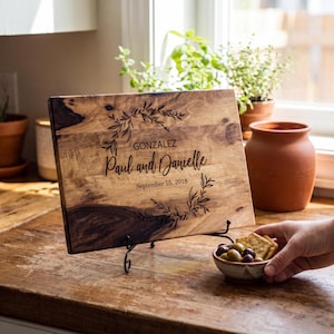 a wooden cutting board with a personalized message engraved on it, placed on a wooden table. A hand is holding a small bowl of olives in the foreground.