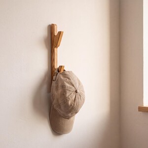 Könnte beinhalten: Eine hellbraune, an der Wand montierte Garderobenleiste aus Holz mit zwei nach oben gerichteten Haken. Eine beige Baseballkappe mit strukturierter Krone und gebogenem Schirm hängt am unteren Haken der an der weißen Wand befestigten Leiste.