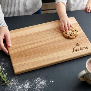 A person is holding a wooden cutting board with a cookie on it, and there is a cup of hot chocolate nearby.