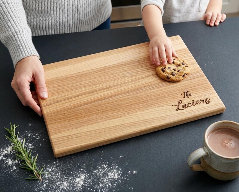 A person is holding a wooden cutting board with a cookie on it, and there is a cup of hot chocolate nearby.