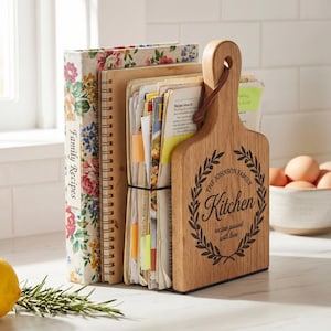 A wooden board with a decorative handle is placed on a kitchen counter, holding a collection of recipe books and a bowl of eggs.