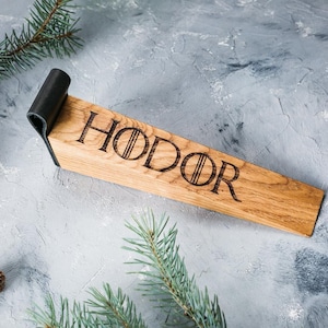 May include: Wooden doorstop with the word "HODOR" etched in black. The doorstop is made of light brown wood and has a black leather handle. The doorstop is rectangular and is resting on a gray surface with pine needles.