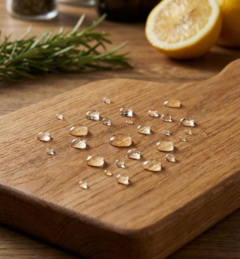 A wooden cutting board with water droplets on it, surrounded by fresh rosemary and a lemon slice.