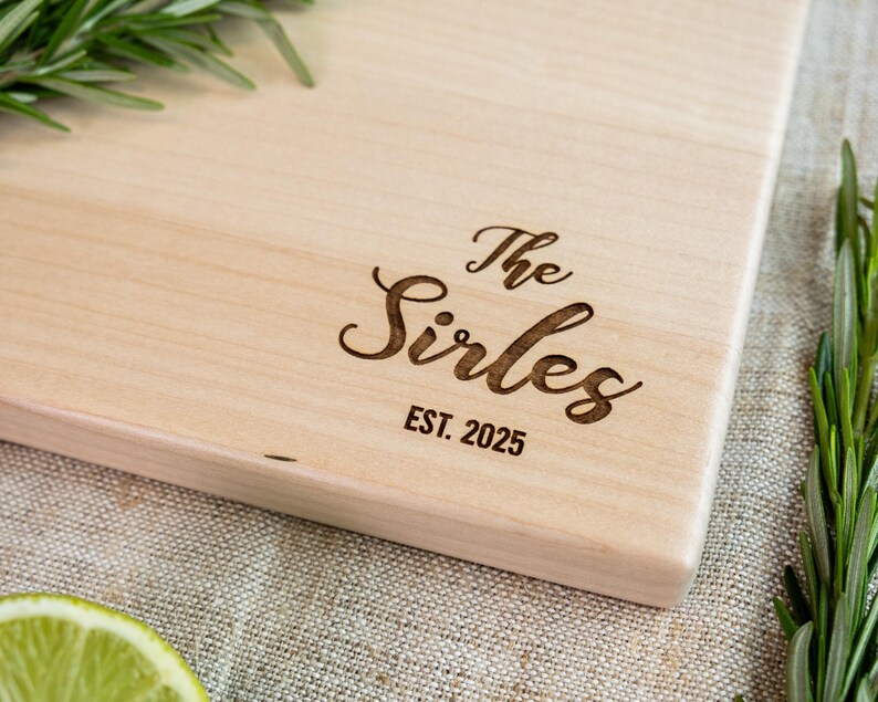 A wooden cutting board with the name "The Sirles" engraved on it, along with the year "EST. 2025" below it.