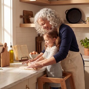 A woman and a young girl are standing in a kitchen, preparing food together. The woman is holding the young girl's hand as they work on a dough or batter, with various kitchen items and ingredients visible in the background.