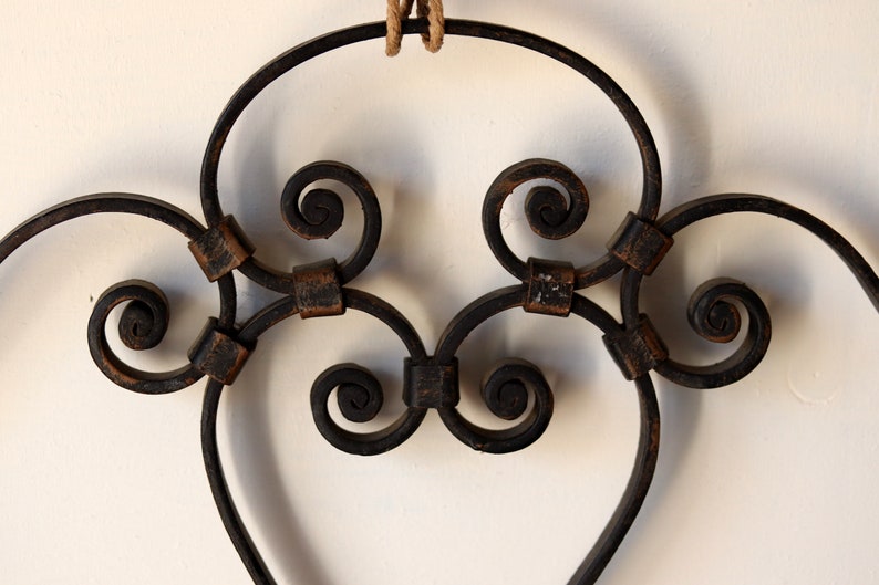 Op de afbeelding: Een zwarte metalen hartvormige wanddecoratie met ingewikkelde krulwerk en een rustieke afwerking.