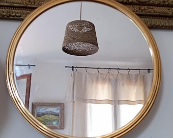 Peut inclure: Un miroir rond avec un cadre doré, reflétant une pièce avec un abat-jour tissé, des rideaux blancs et une peinture de paysage encadrée. Le cadre du miroir présente un design détaillé et nervuré, ajoutant une touche d'élégance vintage.