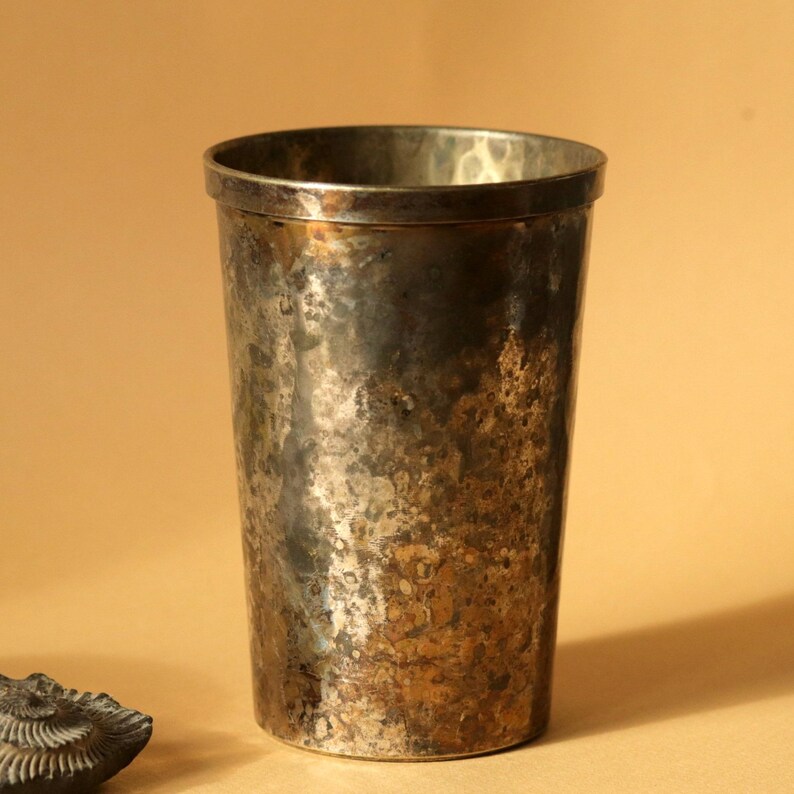 Op de afbeelding: Een hoge metalen beker in antieke stijl met een gehamerde textuur. De beker heeft een donkere, aangetaste uitstraling met brons- en zilvertinten. De rand is glad en gepolijst. De beker wordt waarschijnlijk gebruikt om uit te drinken.
