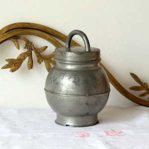 May include: A silver pewter jar with a lid and a handle. The jar has a round body and a slightly flared base. The jar is sitting on a white tablecloth.