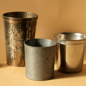 Op de afbeelding: Drie metalen drinkbekers van verschillende maten en afwerkingen worden getoond. De hoogste beker heeft een gehamerde textuur, terwijl de andere glad zijn. De bekers zijn gemaakt van zilver en tin.