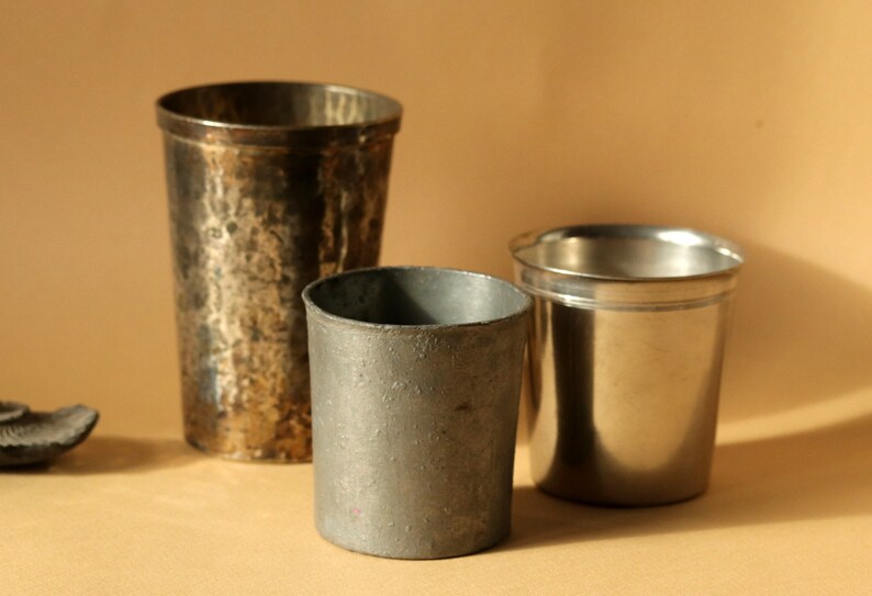 Op de afbeelding: Drie metalen drinkbekers van verschillende maten en afwerkingen worden getoond. De hoogste beker heeft een gehamerde textuur, terwijl de andere glad zijn. De bekers zijn gemaakt van zilver en tin.